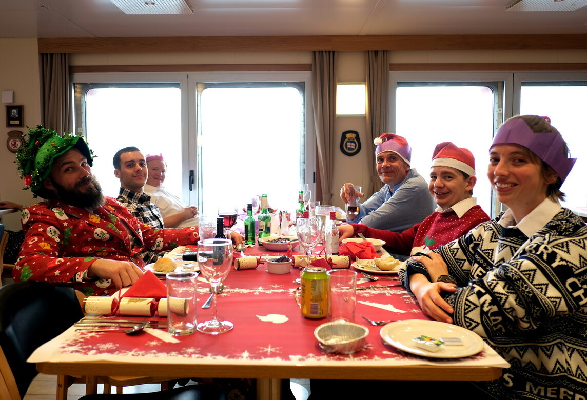 A group of people in Santa hats and paper hats sitting around a long table. Everyone is looking at the camera and smiling. There are crackers and plates and wine glasses on the table