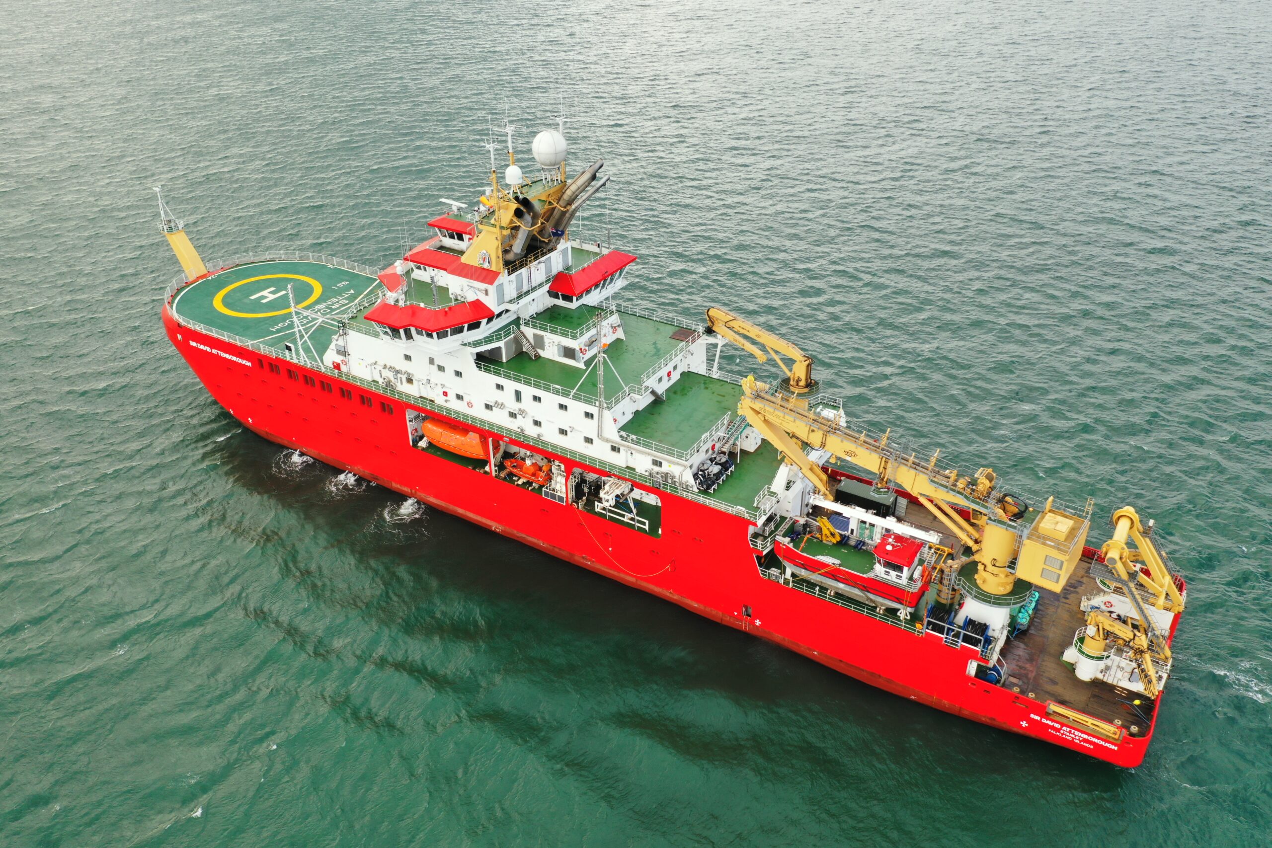 Drone image of a large red ship with several yellow cranes on its deck, and a small boat on its side